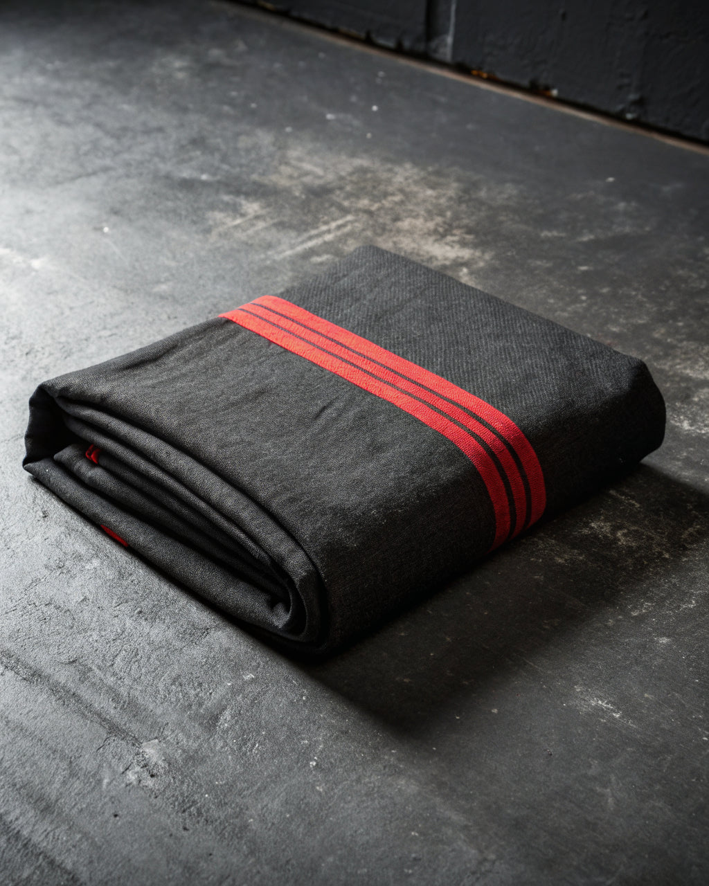 Folded charcoal canvas textile with a single red stripe on dark concrete